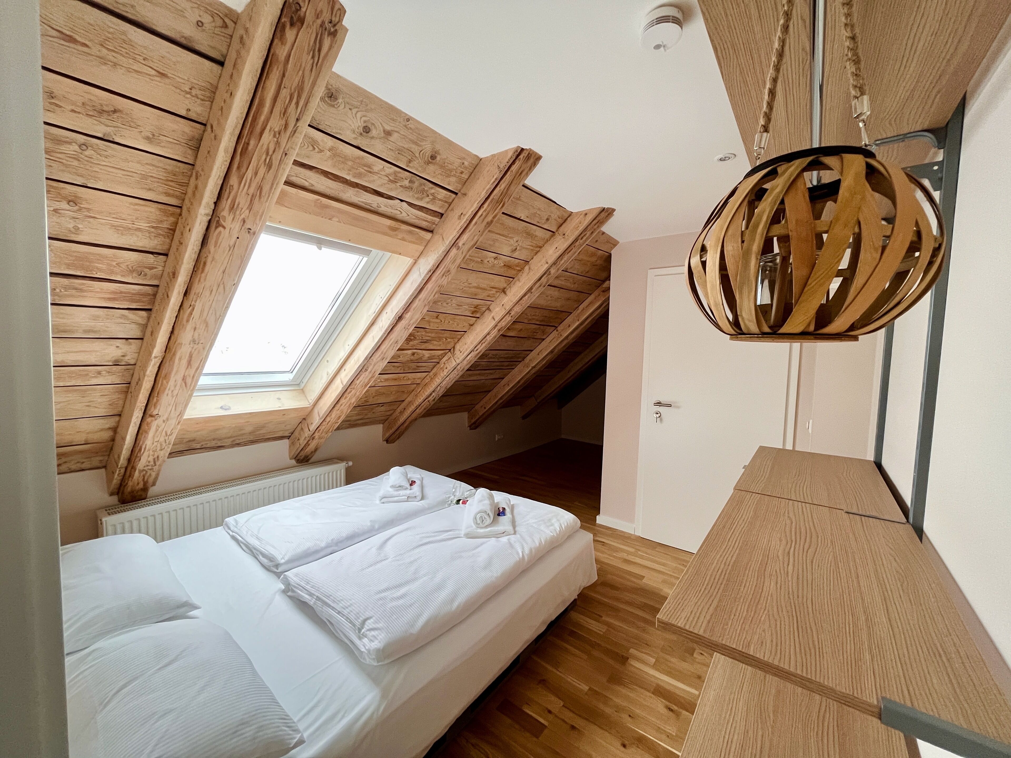 A bedroom with a sloped wooden ceiling, a skylight, a bed with white linens, and a wooden bedside table.
