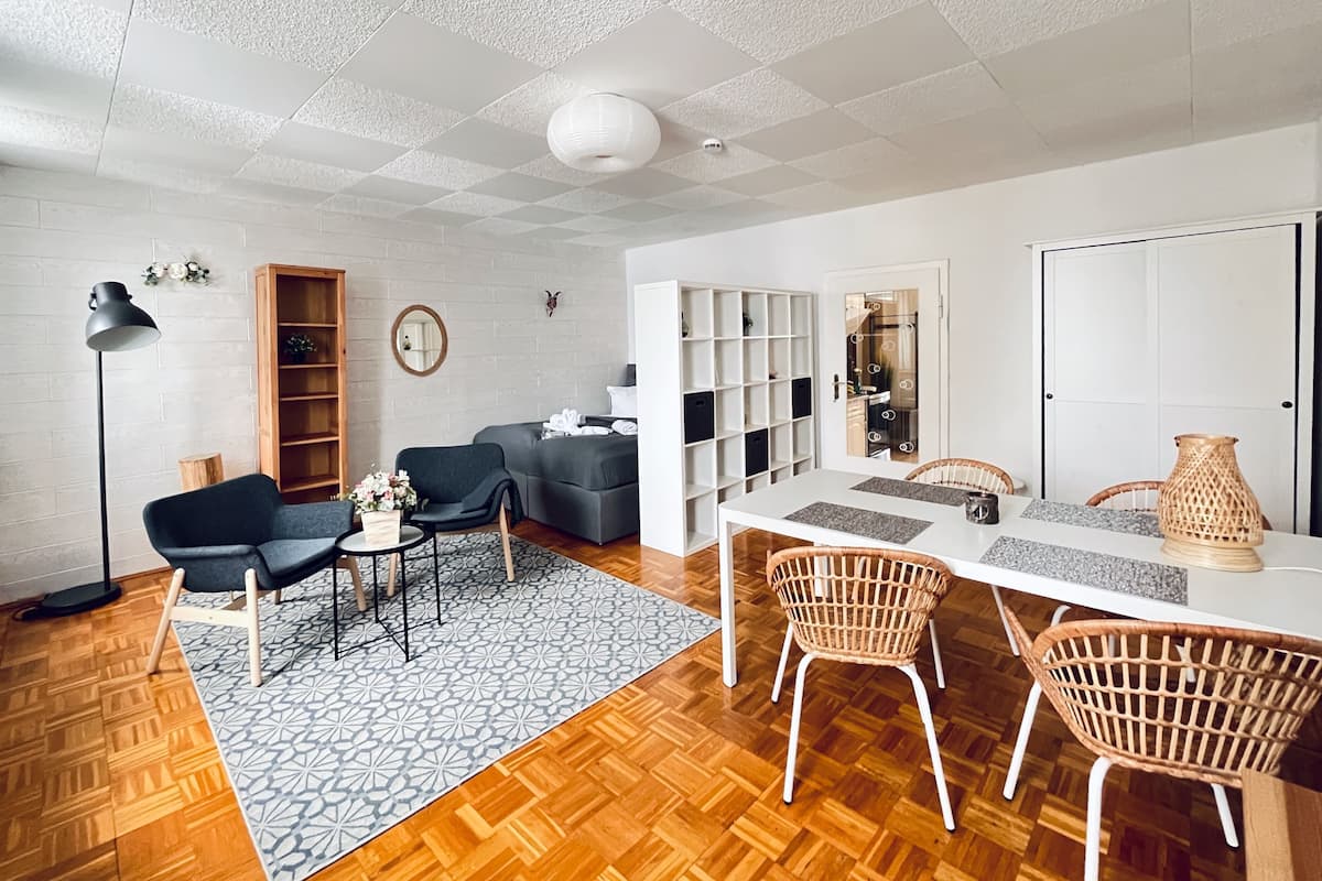 Quarto de hotel moderno com piso de madeira, sofá cinza, mesa de jantar com cadeiras de vime e teto branco com padrão texturizado.