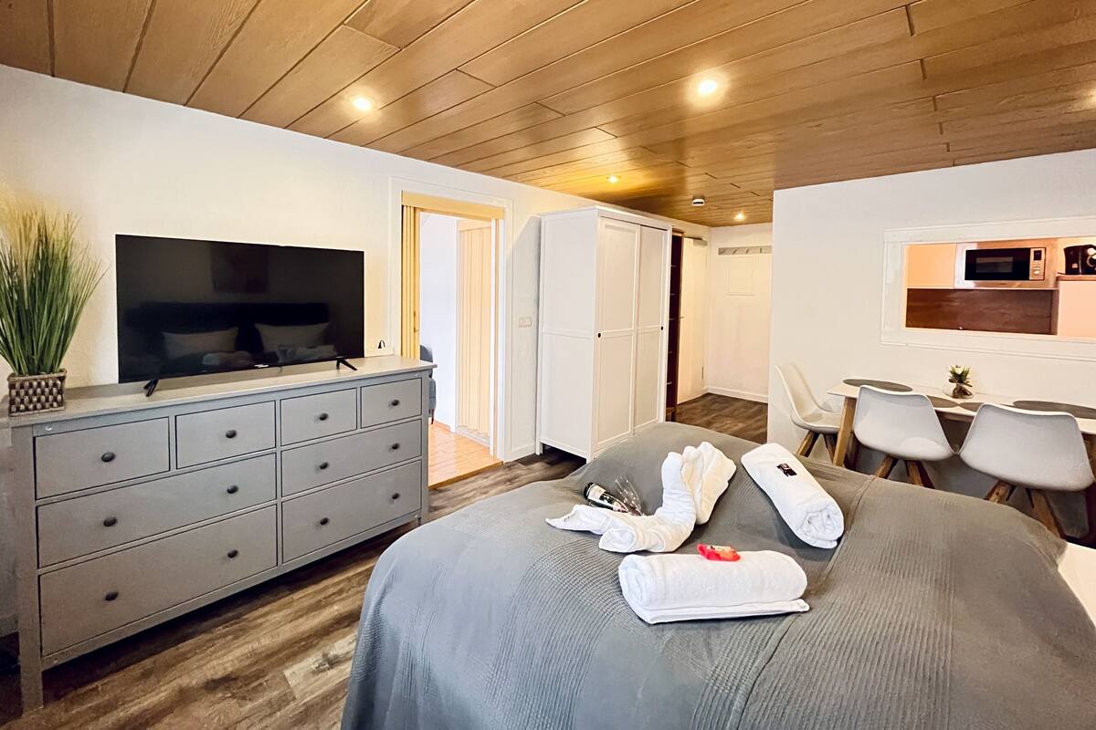 Quarto de hotel moderno com teto de madeira, cama cinzenta com toalhas, televisão em uma cômoda e uma kitchenette.