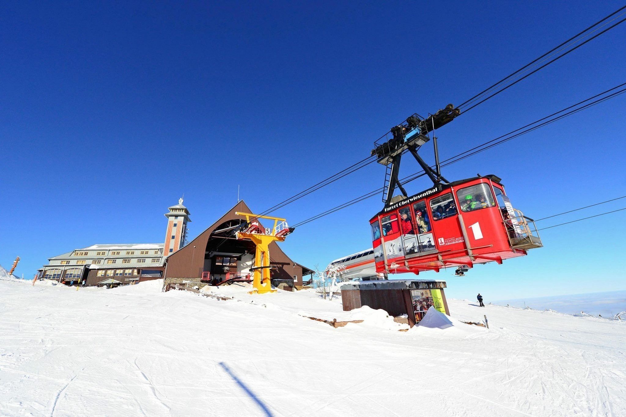 A ski lift with a red cabin and a yellow chairlift, a building with a tower, and a snowy landscape.