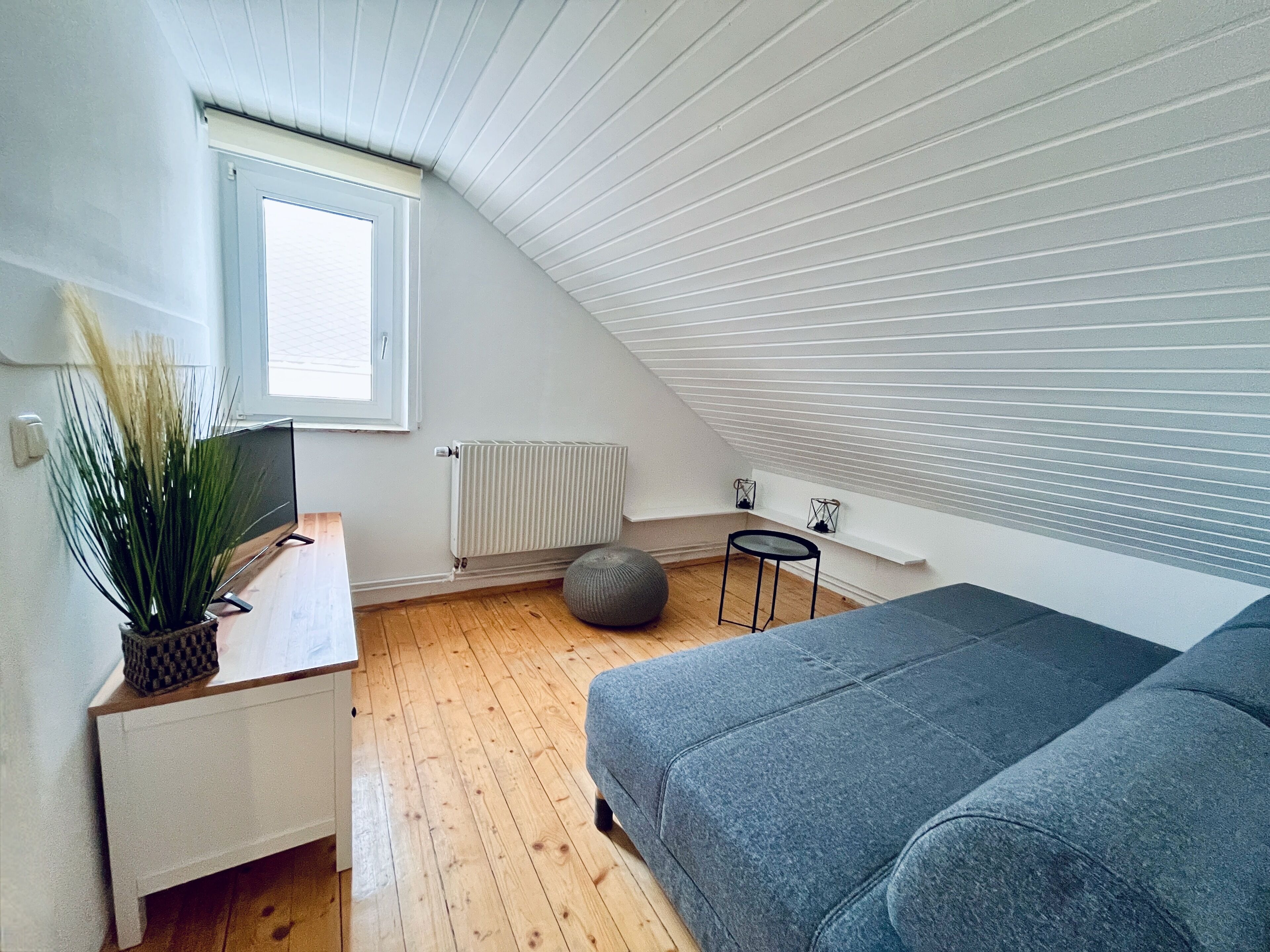 A modern loft bedroom with a sloped ceiling, wooden flooring, a grey sofa, a small round ottoman, a white bedside table with a plant, a radiator, and a window with blinds.