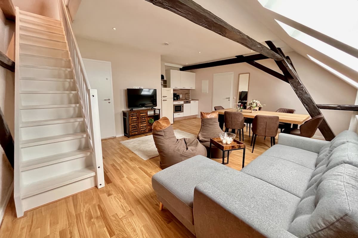 Um apartamento loft moderno com escada, sala de estar com sofá e poltronas, área de jantar com mesa e cadeiras e cozinha ao fundo.