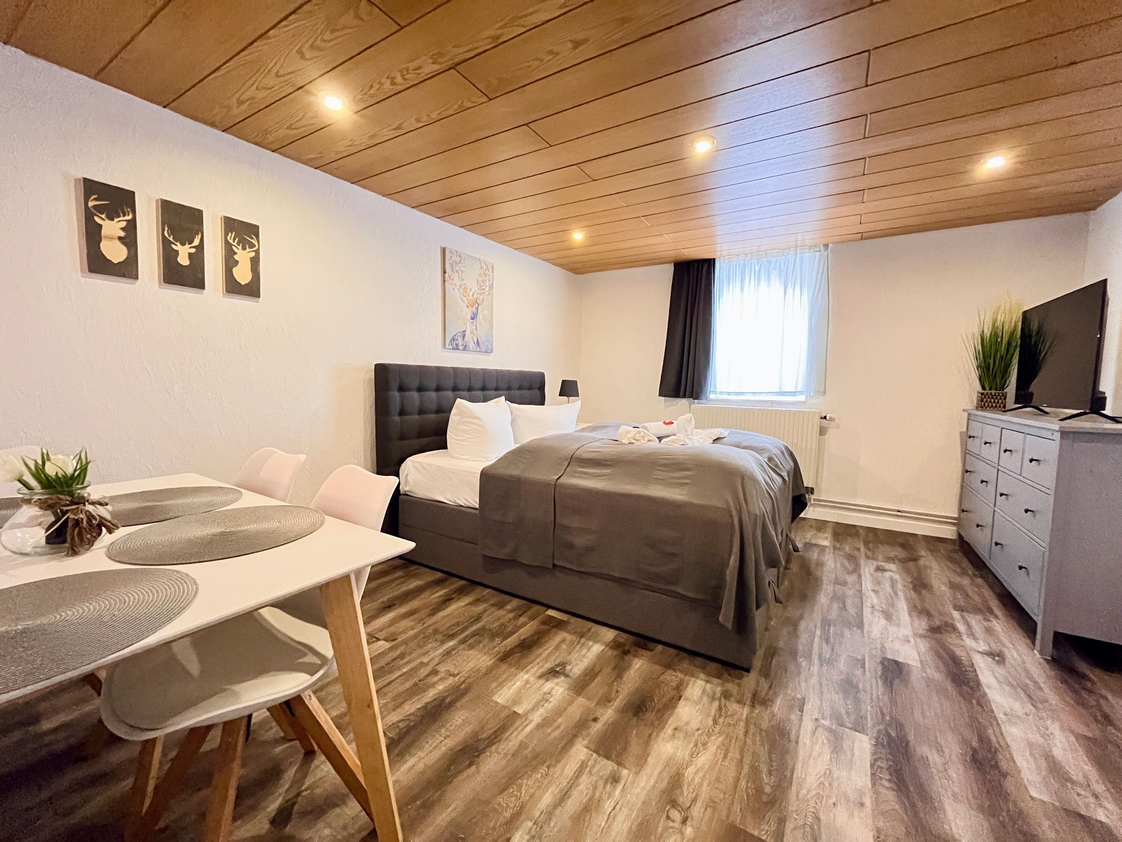 A modern bedroom with a wooden ceiling, a bed with grey bedding, a desk with a chair, a dresser, and a television on the dresser.