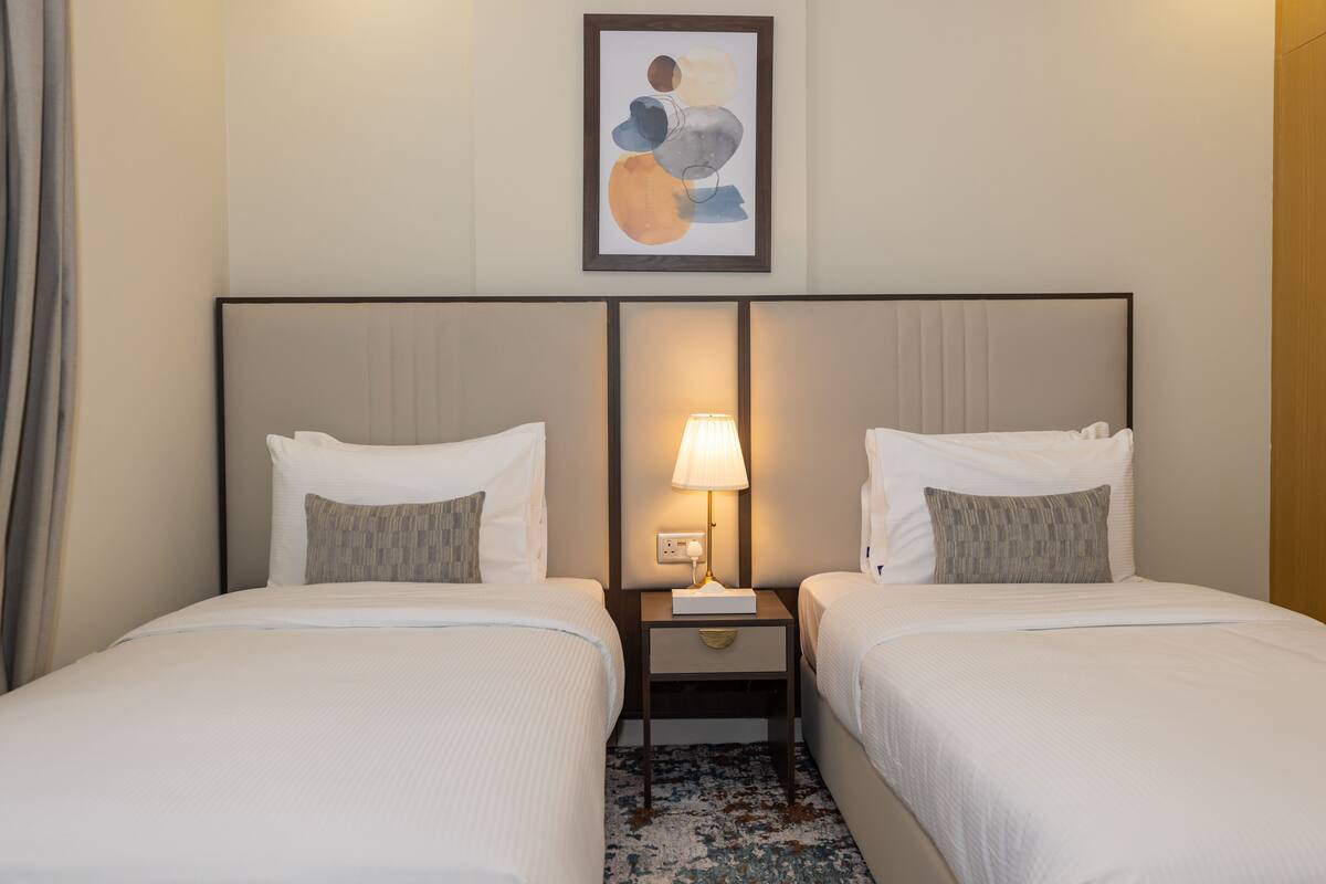 Une chambre d’hôtel avec deux lits, une table de chevet avec une lampe, une œuvre d’art abstraite encadrée au mur et un tapis à motifs.