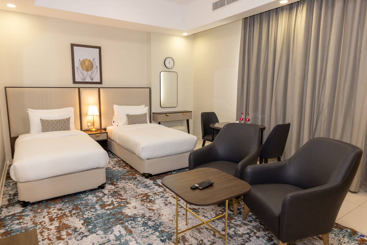Une chambre d’hôtel avec deux lits, un bureau, une chaise, une petite table, une horloge et un tableau encadré au mur.