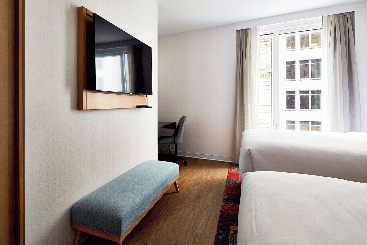 A hotel room with a bed, a bench, a desk, a chair, a TV, and a window with curtains.