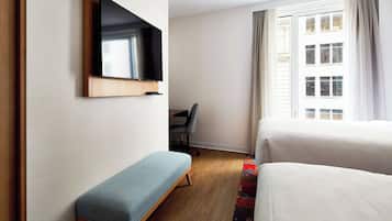 Quarto de hotel com cama, um banquinho, uma cadeira, televisão fixada na parede e janela com cortinas.