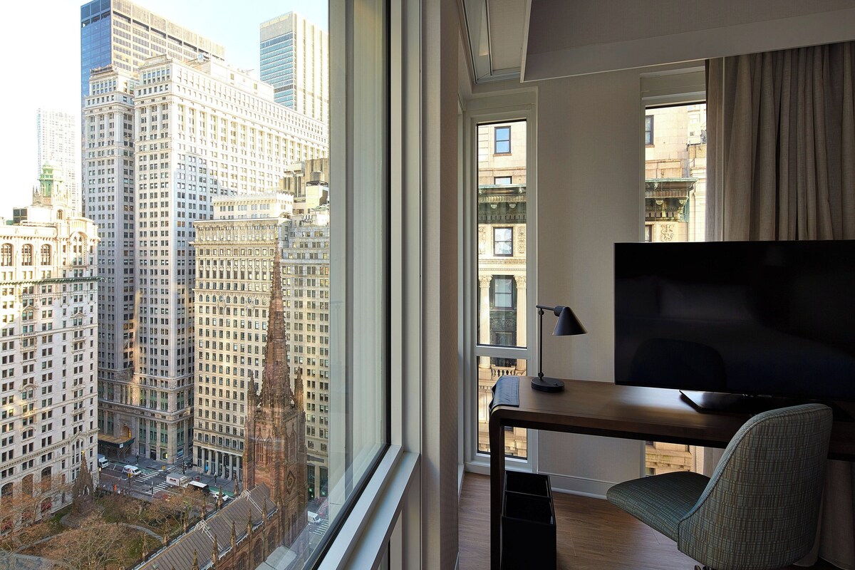 A hotel room with a large window offering a city view, a flat-screen TV, and a desk with a chair.