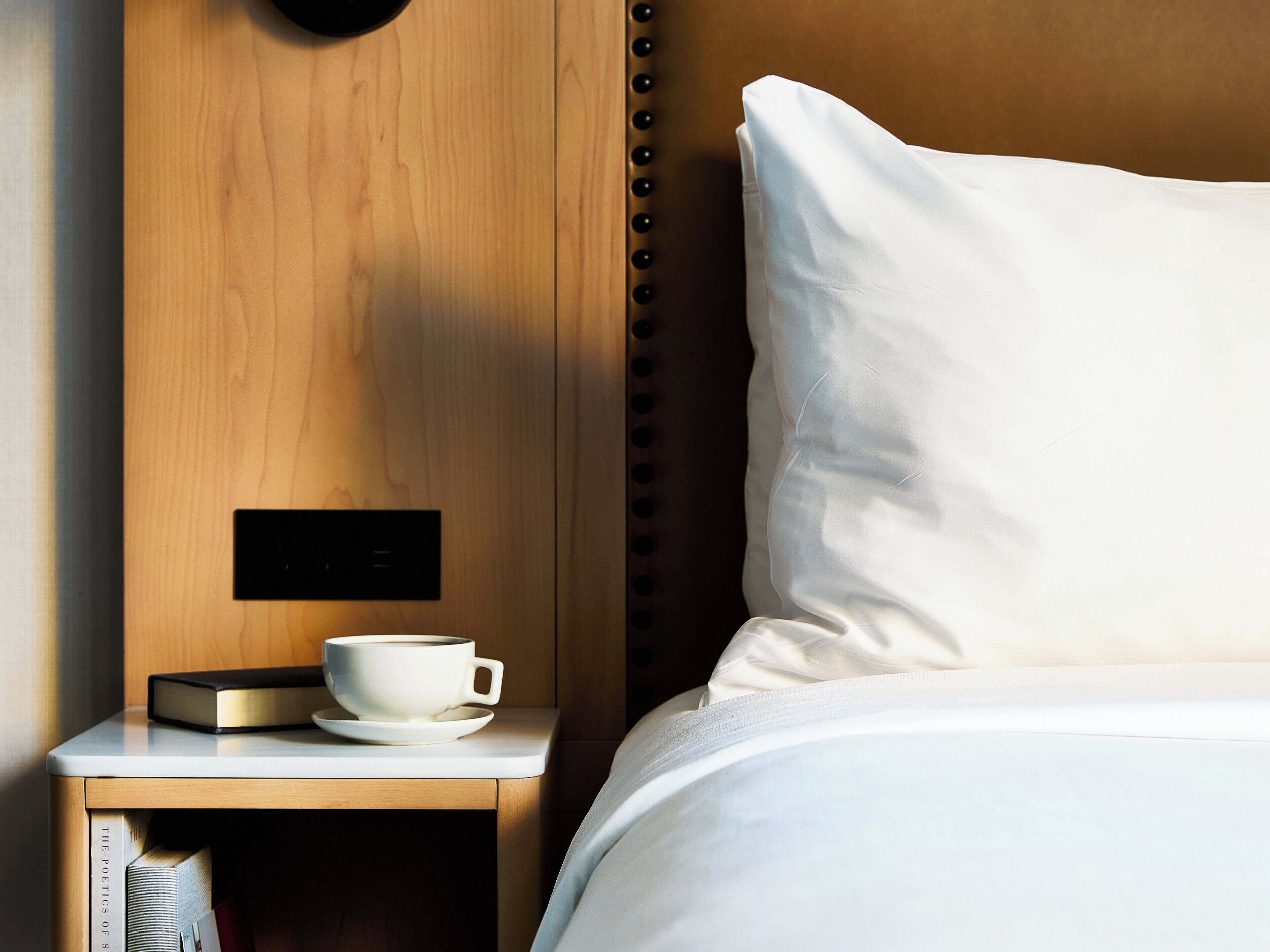 A neatly made bed with a white pillow and a wooden nightstand beside it. On the nightstand, there is a white cup and saucer, and a book titled "The Poetics of Sleep".