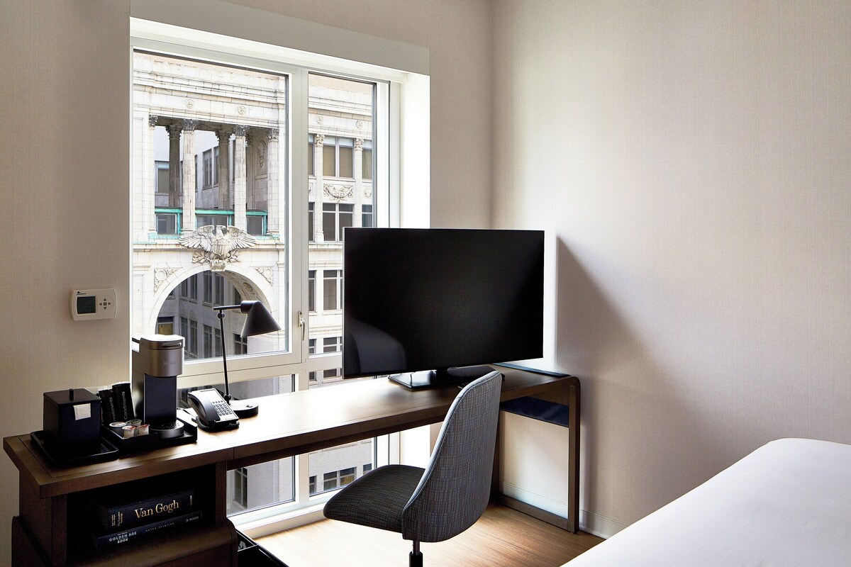 A hotel room with a large window, a desk with a flat-screen TV, a chair, and a bed.
