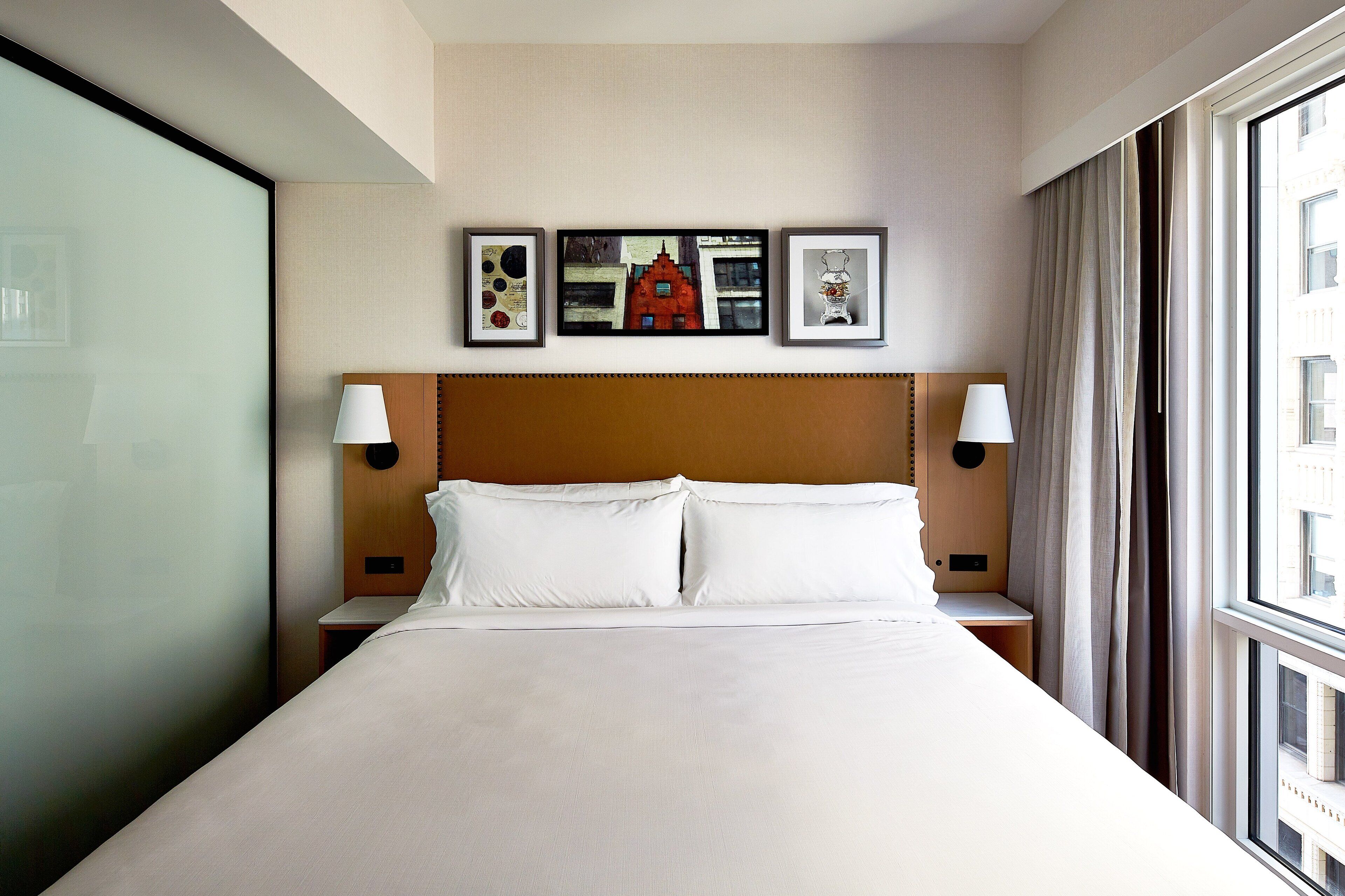 Una habitación de hotel con una cama grande, un cabecero con dos lámparas fijadas a la pared y tres cuadros enmarcados.