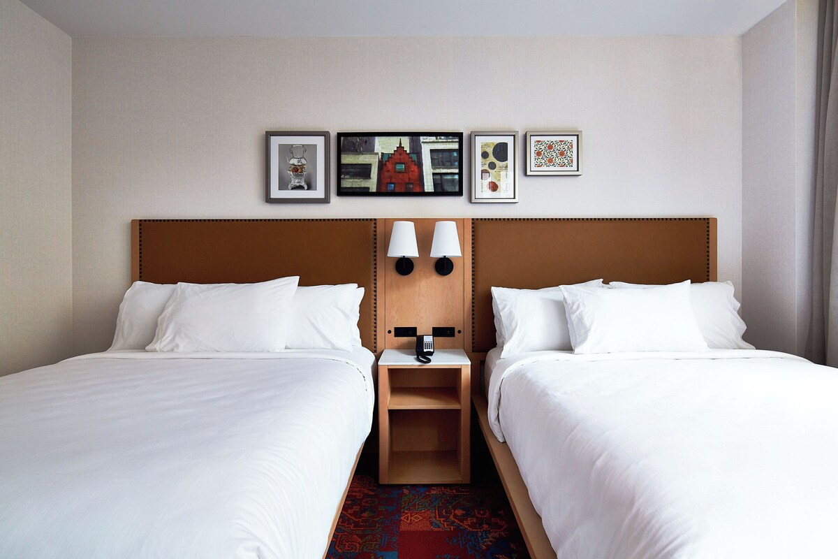 A hotel room with two single beds, a wooden headboard, a nightstand with a telephone, and a wall with framed artwork.
