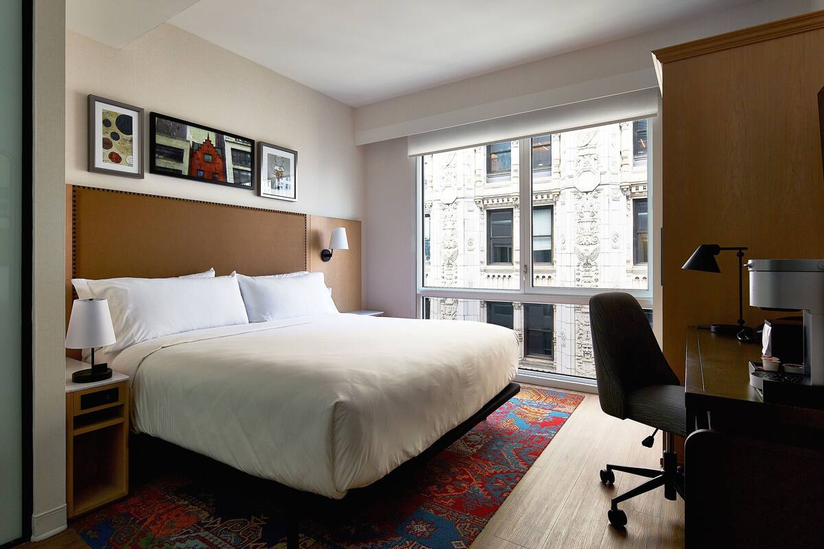 Une chambre d’hôtel avec un grand lit, un bureau avec une chaise, une fenêtre donnant sur un immeuble et des tableaux encadrés accrochés au mur.