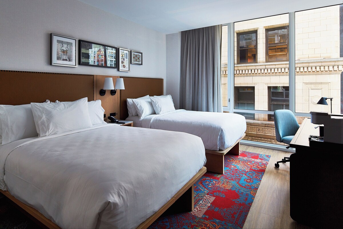 A hotel room with two beds, a desk, a chair, and a large window with a view of buildings.
