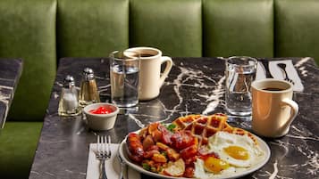 Ein Frühstücksteller mit Wurst, Kartoffeln und einem Spiegelei, dazu Kaffee und Beilagen auf einer Marmortischplatte.