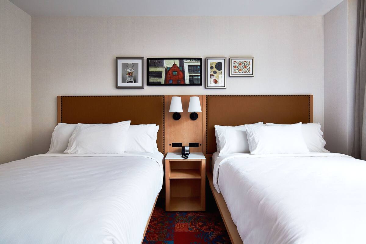 Une chambre d’hôtel avec deux lits simples, une tête de lit en bois, une table de chevet avec un téléphone et un mur orné de tableaux.