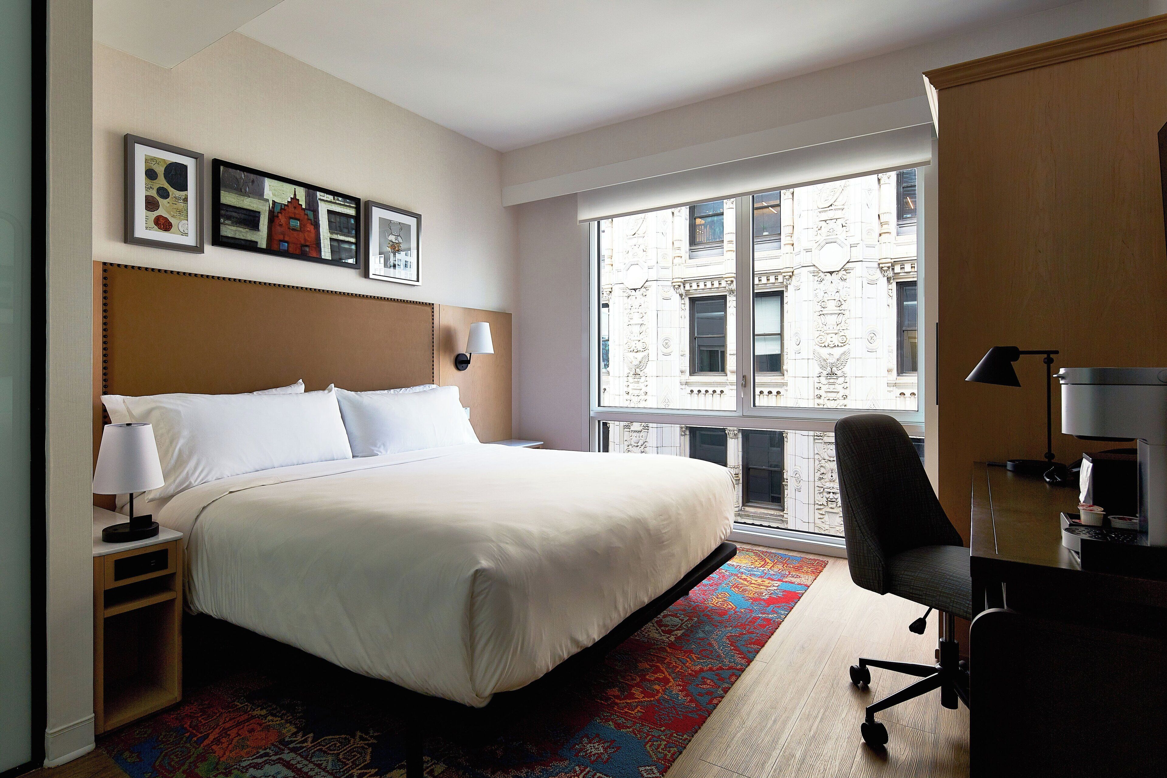 A hotel room with a large bed, a desk with a chair, a window with a view of buildings, and artwork on the wall.
