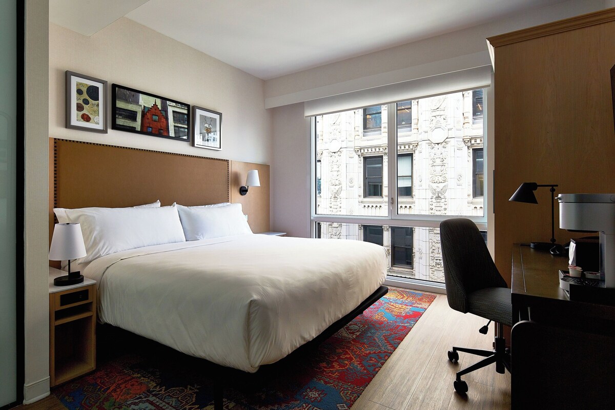 A hotel room with a large bed, a desk with a chair, a window with a view of a building, and framed pictures on the wall.