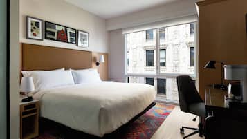 Een hotelkamer met een groot bed, een bureau met stoel, een raam met uitzicht op gebouwen en kunstwerken aan de muur.