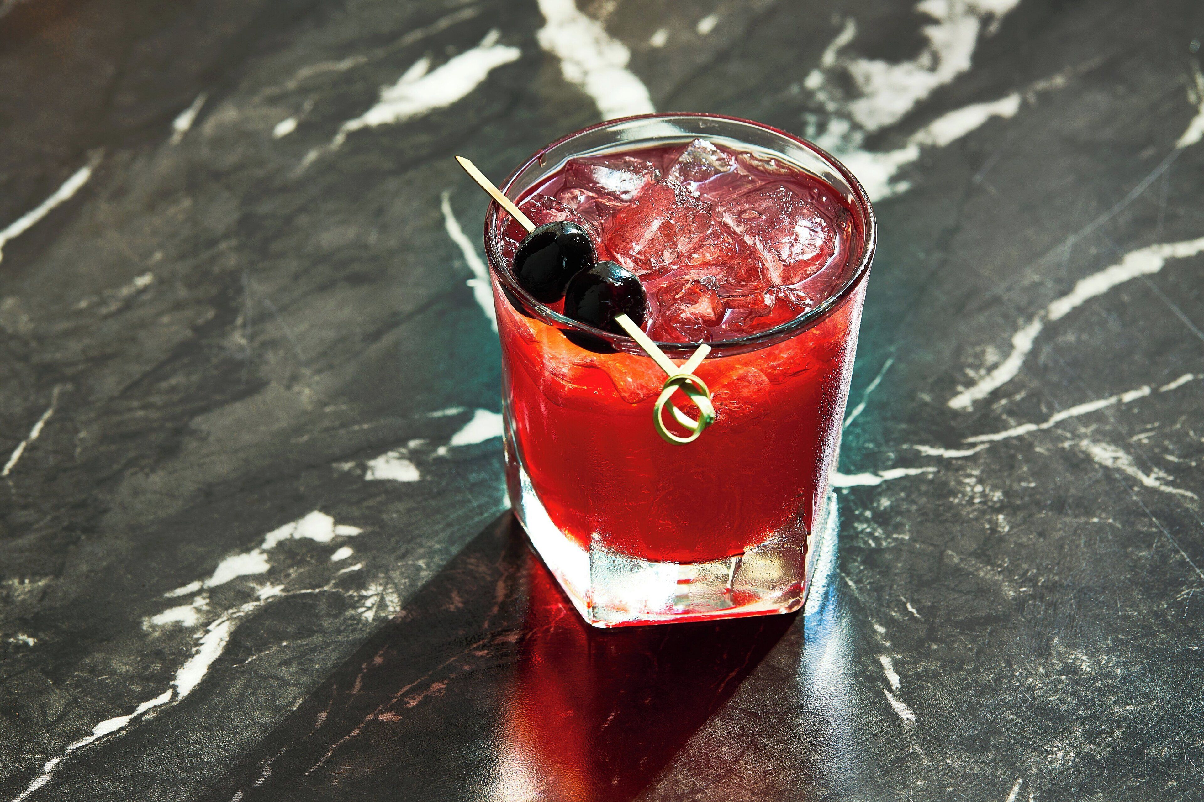 A glass of red beverage with ice cubes and two olives on a dark marble surface.