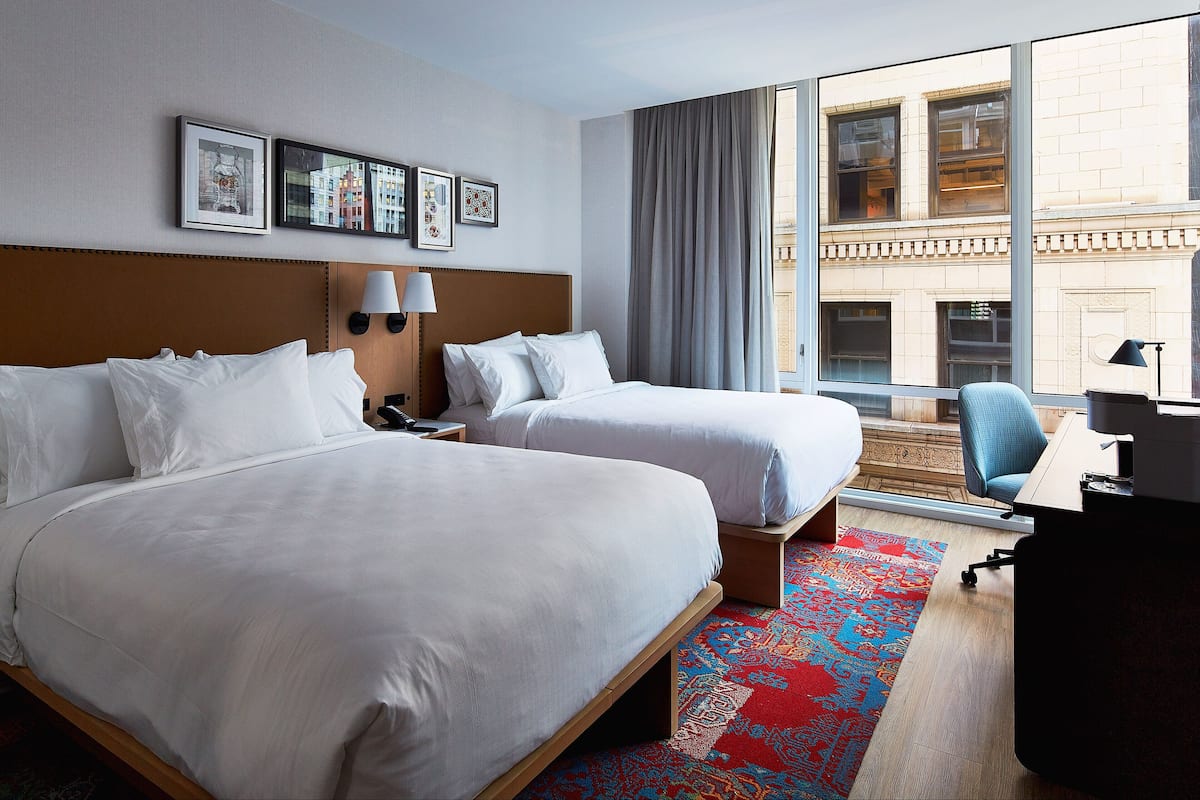 Une chambre d’hôtel avec deux lits, un bureau, une chaise et une grande fenêtre donnant sur des immeubles.