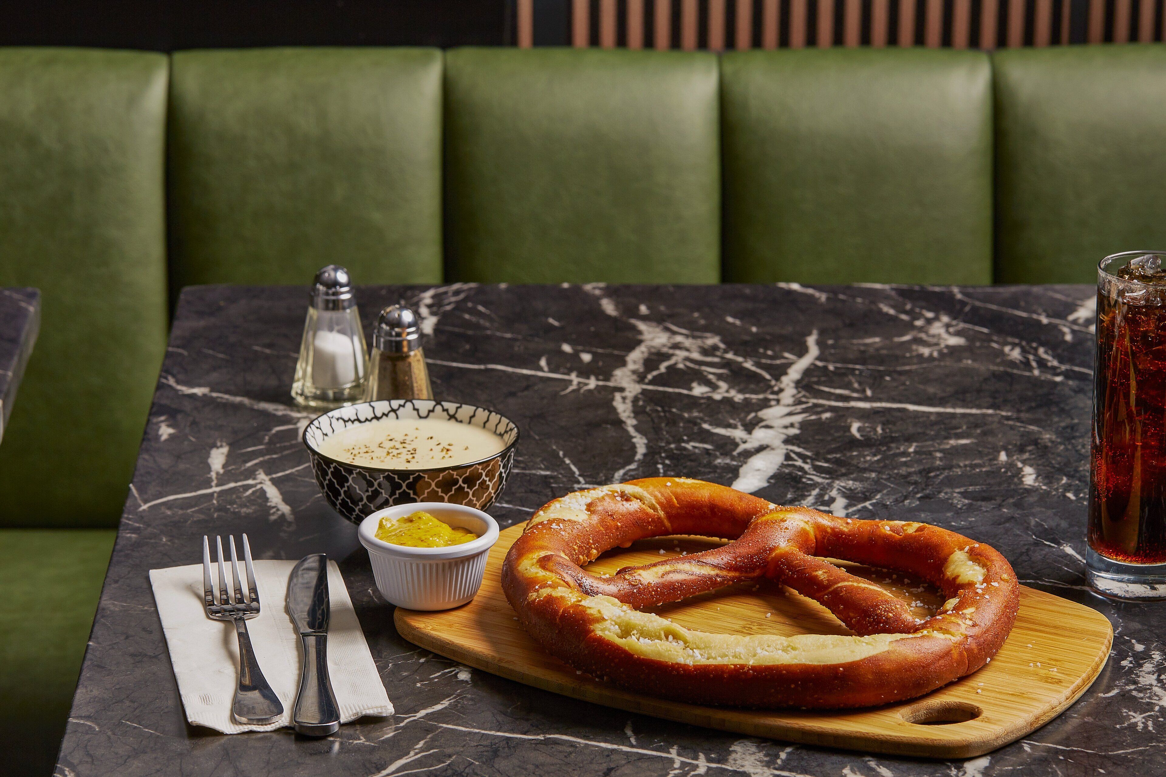 A dining table with a pretzel, a bowl of mustard, and a glass of iced tea.