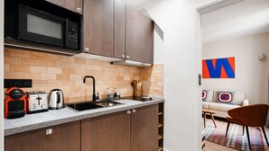 Eine moderne Küche mit dunklen Holzschränken, einer Mikrowelle, einem Toaster und einem Wasserkocher. Ein Spülbecken ist zu sehen, und im angrenzenden Wohnbereich liegt ein farbenfroher Teppich.