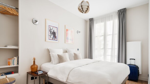 Ein Hotelzimmer mit einem großen Bett, einem Nachttisch, einer Lampe, einem Fenster mit Vorhängen und einem Bücherregal.
