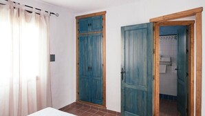 A bedroom with a bed, a window with sheer curtains, and two doors leading to a bathroom.