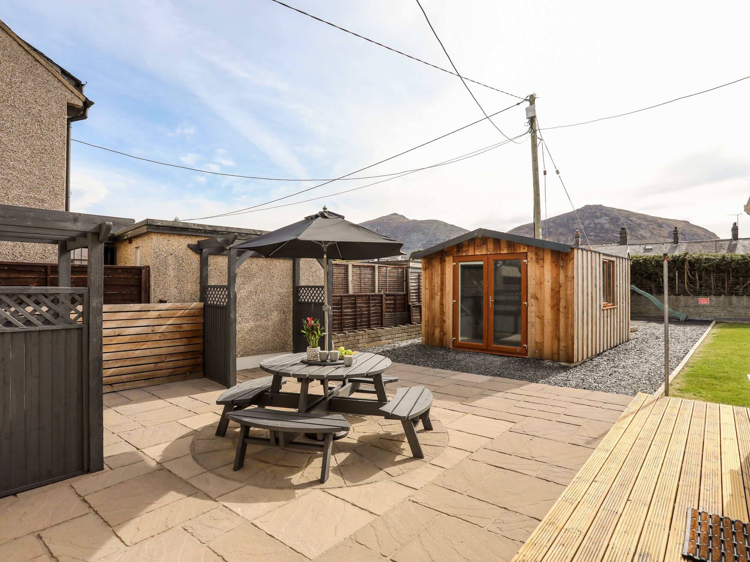 Eine Terrasse mit einem Holztisch und Bänken, eine kleine Holzkonstruktion mit Glastüren und ein Holzzaun.