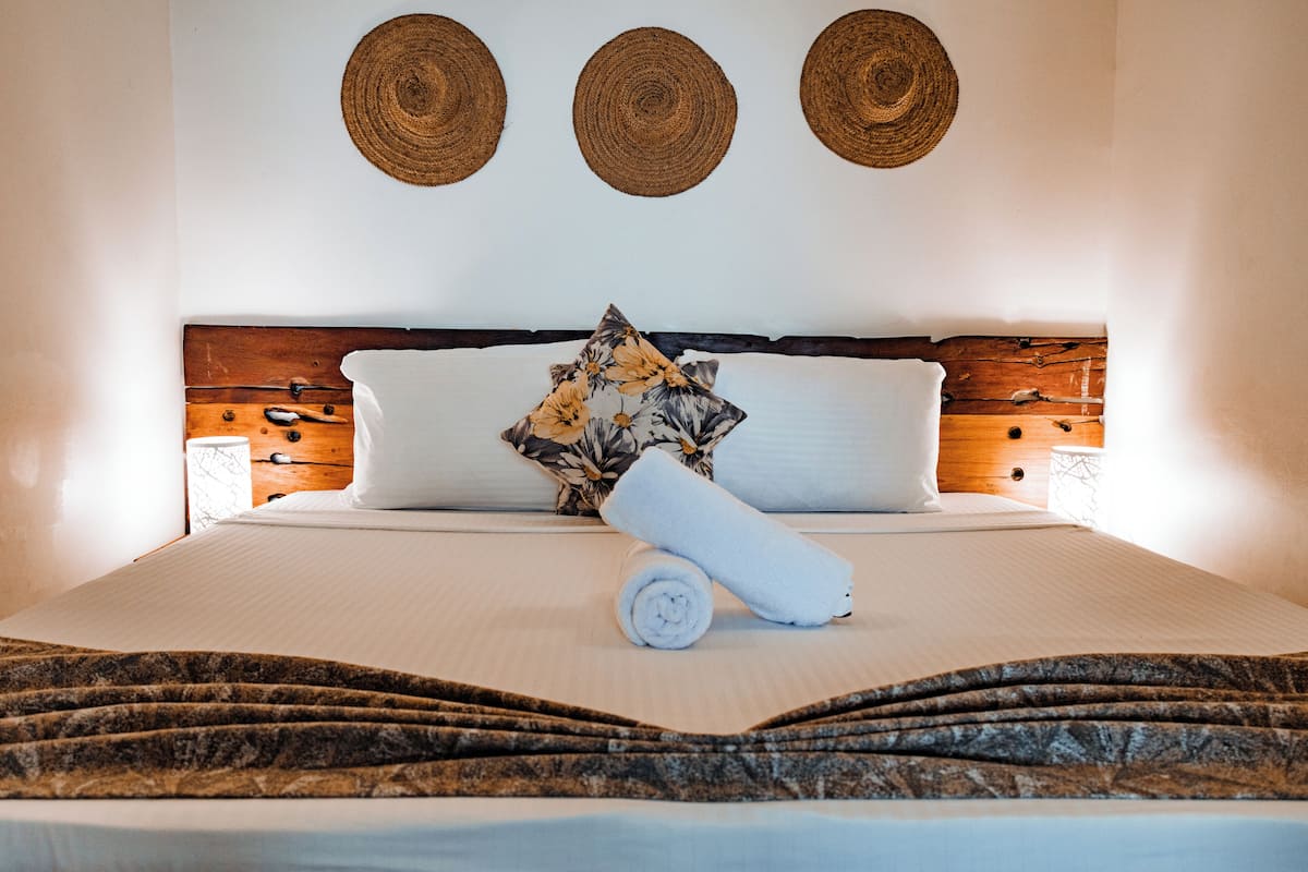Una cama bien tendida con una toalla enrollada, un cojín floral y un cabecero de madera.