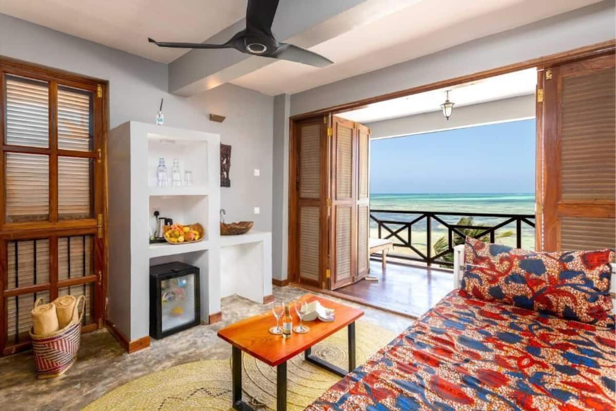 Una habitación con cama, una mesa con vasos, una estantería con botellas y vistas al océano.