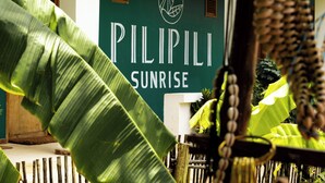 Letrero de 'Pilipili Sunrise' con fondo tropical que incluye hojas de plátano y una valla de madera.