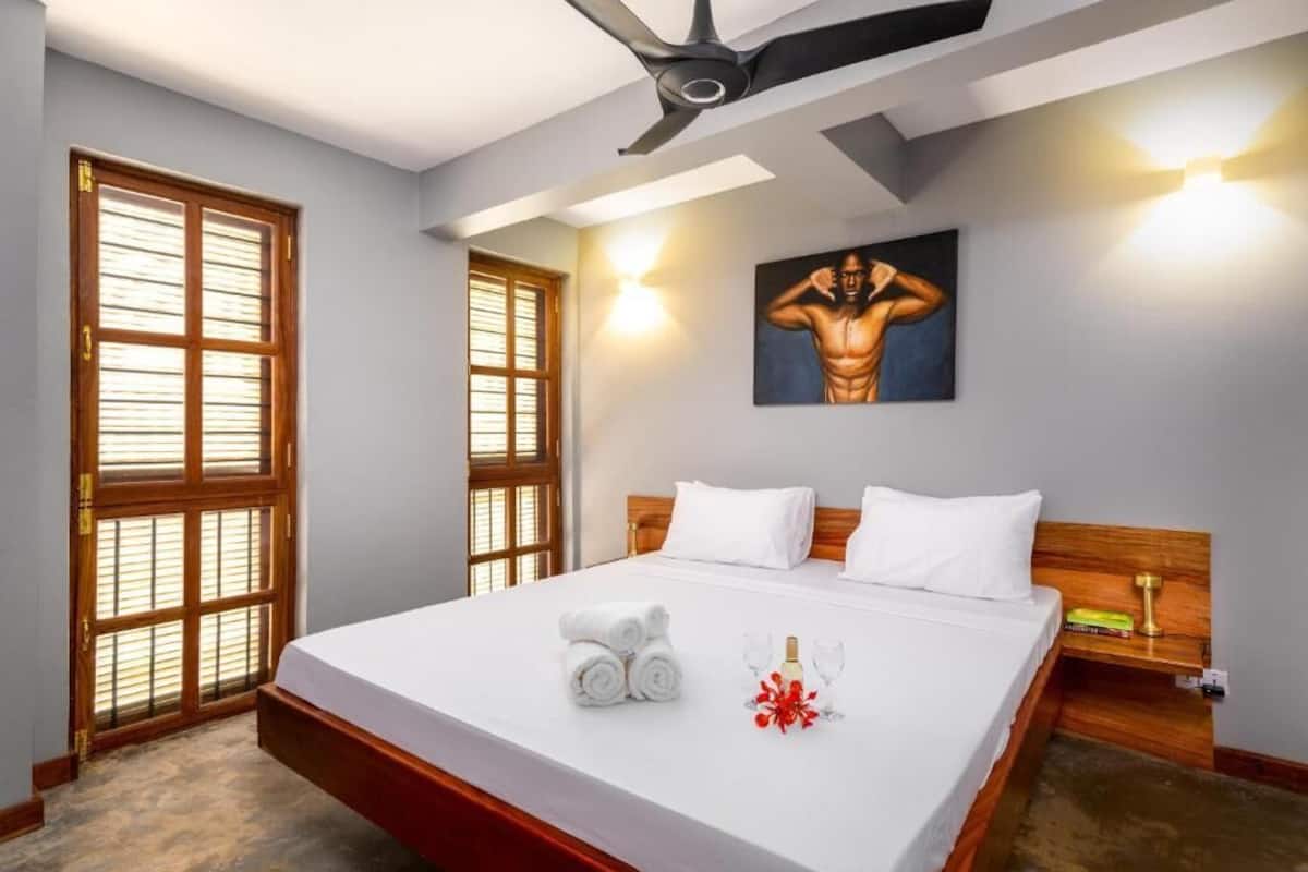 Una habitación de hotel con una cama grande, dos ventanas con persianas, un ventilador de techo, un cuadro en la pared y una mesita de noche con una botella y vasos.