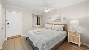 A bedroom with a bed, a nightstand, a ceiling fan, a window with curtains, and a door. - Oceanfront Sunrises in Upgraded Seaside Villa 225 (Hilton Head Island)