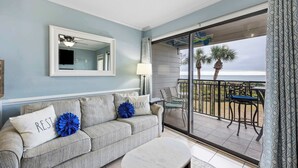 A living room with a grey sofa, a coffee table, a ceiling fan, a large window with a view of palm trees, and a balcony with outdoor furniture. - Oceanfront Sunrises in Upgraded Seaside Villa 225 (Hilton Head Island)