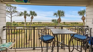 A balcony with a table and chairs, overlooking a beach with palm trees and a clear sky. - Oceanfront Sunrises in Upgraded Seaside Villa 225 (Hilton Head Island)