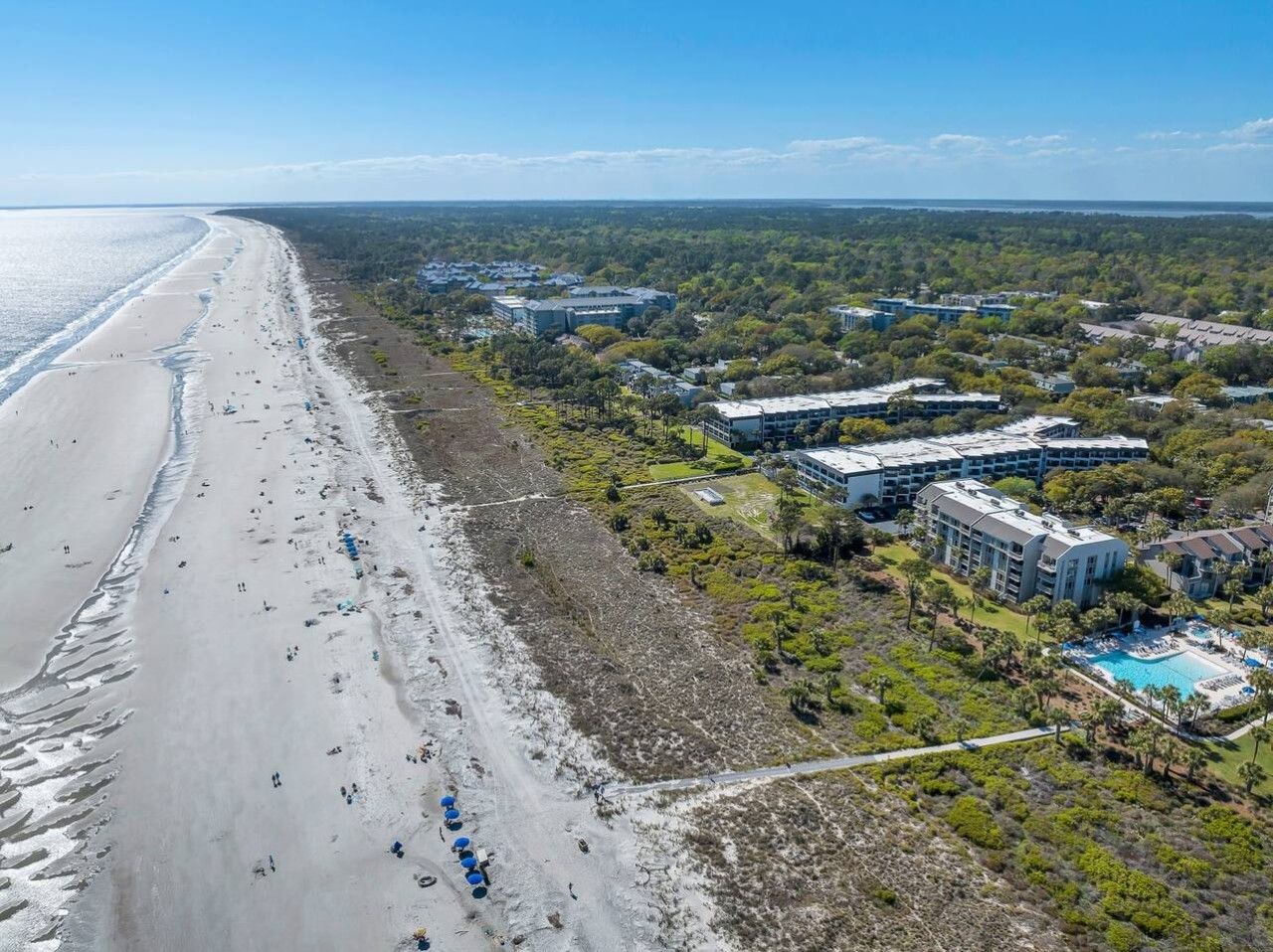 Vista aérea de una zona costera con una playa, un complejo turístico con varias edificaciones y una piscina.