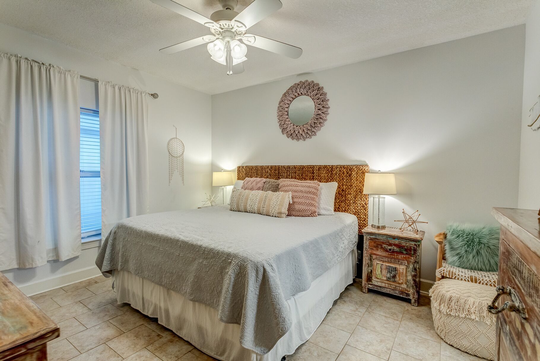 A bedroom with a bed, bedside lamps, a decorative wall mirror, a ceiling fan, and a window with curtains.