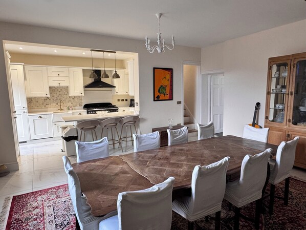 Un coin repas avec une grande table en bois et des chaises blanches, une cuisine en arrière-plan et un lustre.
