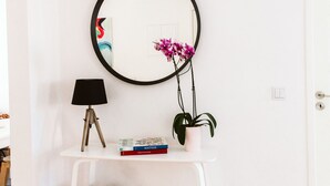 A white wall with a round mirror, a small white table, a lamp, a potted plant, and a few books. - NEW! Belém Riverside 2 bedrooms completely renovated (Lisboa)