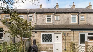 A stone house with a small patio, a white door, and a window with a metal grill. - Magdala (Buxton)