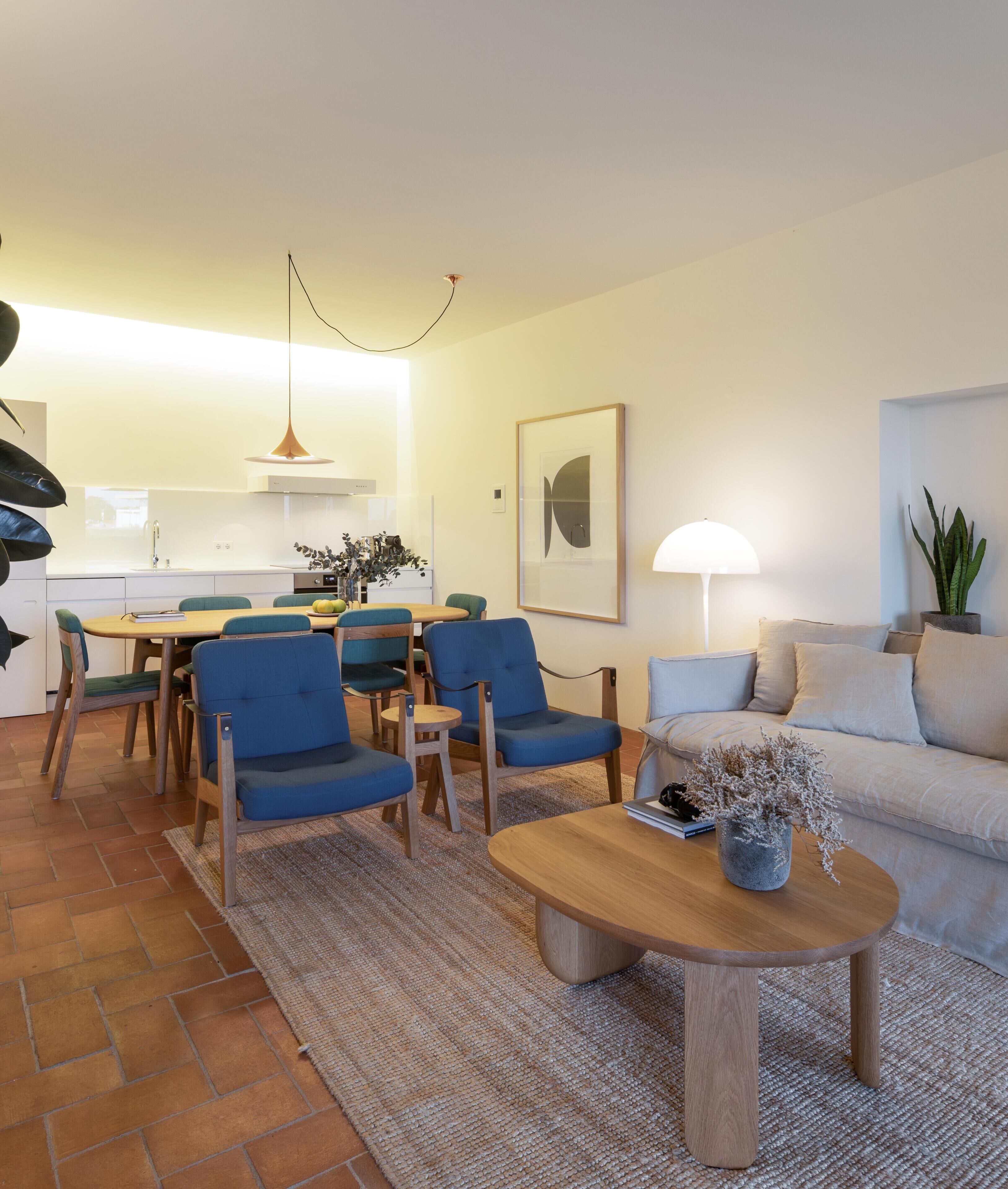 A modern living room with a wooden coffee table, blue chairs, a beige sofa, and a dining area with a table and chairs.