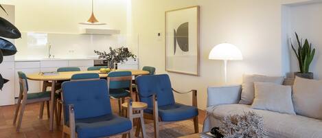 A modern living room with a wooden coffee table, blue chairs, a beige sofa, and a dining area with a table and chairs.