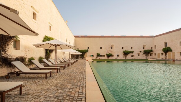 Zona de piscina con sillones de descanso y sombrillas, un sendero pavimentado en piedra y un edificio con tejado de azulejos.