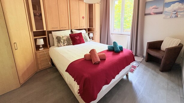 Een slaapkamer met een bed, een houten garderobe, een raam met gordijnen, een stoel en een schilderij aan de muur.