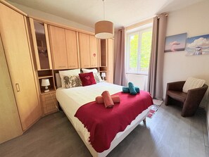 Ein Schlafzimmer mit Bett, Holzschrank, einem Fenster mit Vorhängen, einem Sessel und einem Bild an der Wand.