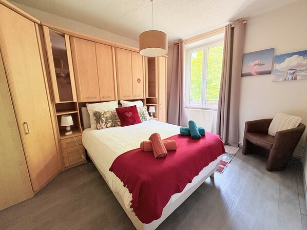 Un dormitorio con cama, armario de madera, ventana con cortinas, una silla y un cuadro en la pared.