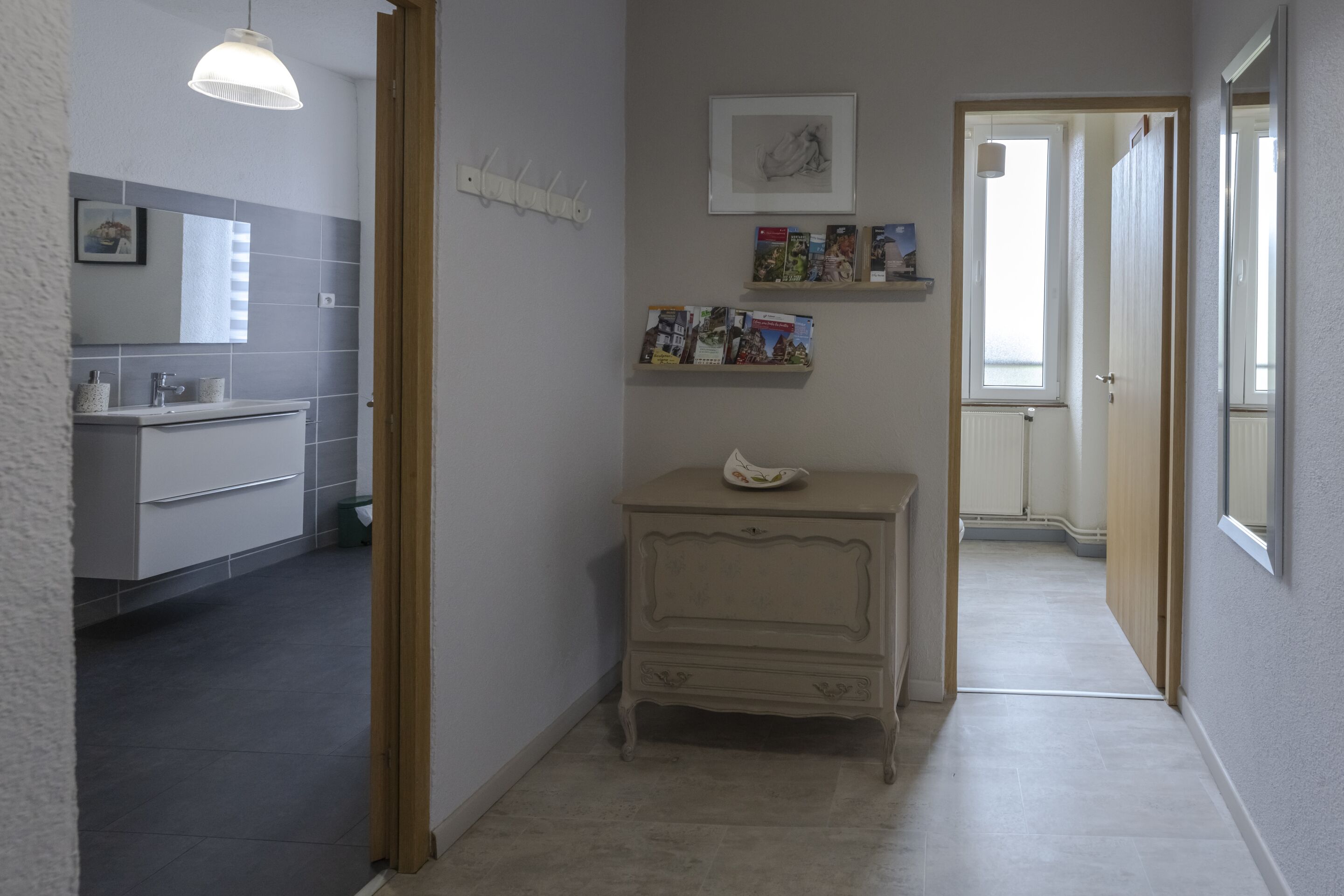 A hallway with a bathroom on the left, a small cabinet with a decorative item, and a door leading to another room.