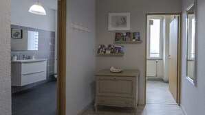 Een gang met aan de linkerkant een badkamer, een klein dressoir met spiegel, en een deur naar een andere kamer.